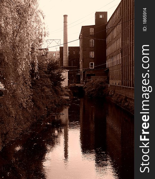Sepia image of canal behind old factory building in easthampton massachusetts, on the left is a bank of trees, on the right a bank of factory windows, in the center a smoke stack. Sepia image of canal behind old factory building in easthampton massachusetts, on the left is a bank of trees, on the right a bank of factory windows, in the center a smoke stack