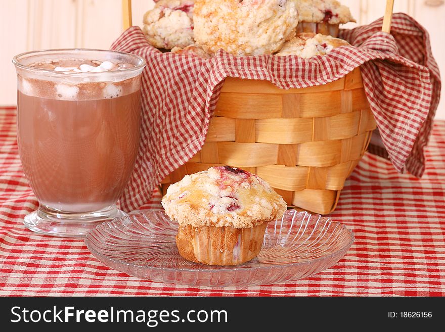 Raspberry muffin on a clear glass plate with a glass mug of hot cocoa and a basket of raspberry muffins on a red checked table cloth. Raspberry muffin on a clear glass plate with a glass mug of hot cocoa and a basket of raspberry muffins on a red checked table cloth