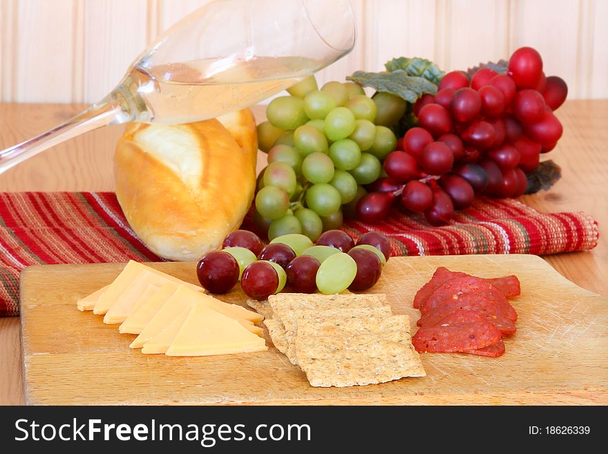 Cheese, crackers, pepperoni, grapes arranged on a cutting board with bread, wine and grapes in the background. Cheese, crackers, pepperoni, grapes arranged on a cutting board with bread, wine and grapes in the background