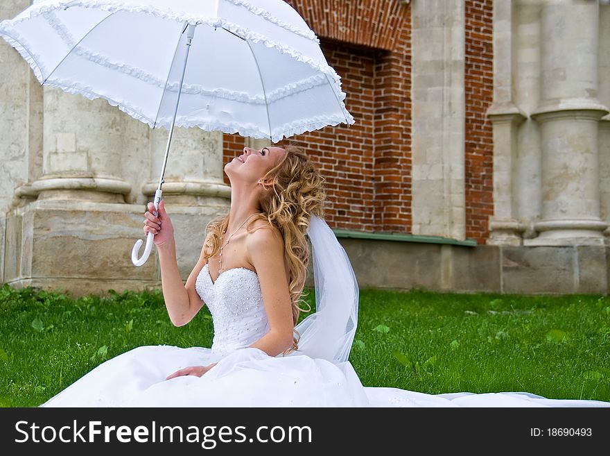 Blonde bride in white dress sitting on the grass at the brick walls of the palace, and looking up at white umbrella. Blonde bride in white dress sitting on the grass at the brick walls of the palace, and looking up at white umbrella