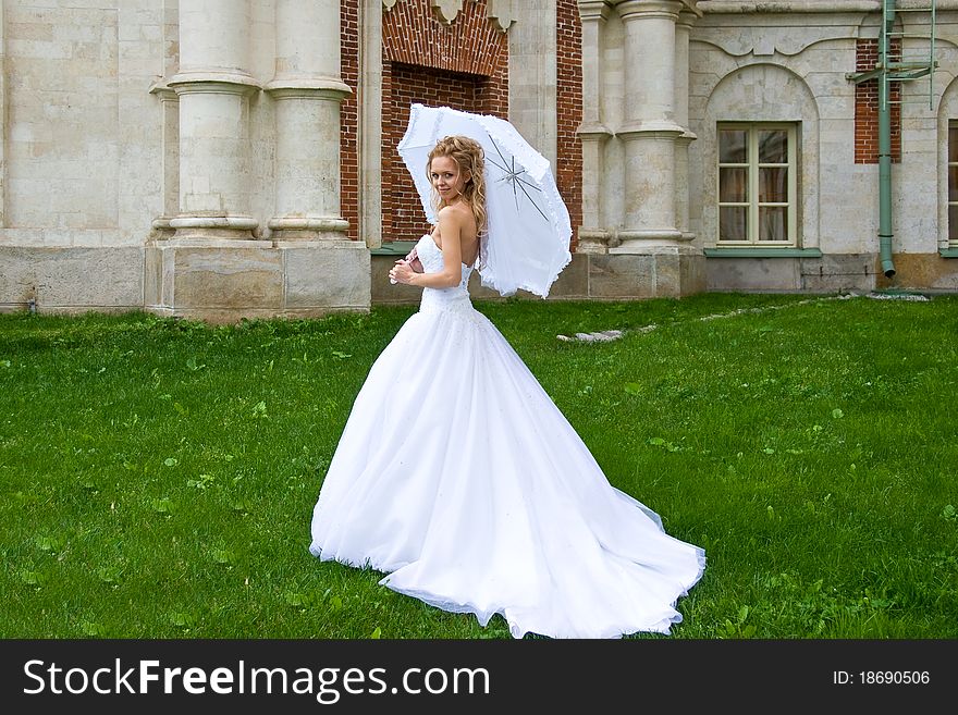 Blonde bride in white dress stands on the grass at the brick walls of the palace under a white umbrella and looking at us. Blonde bride in white dress stands on the grass at the brick walls of the palace under a white umbrella and looking at us