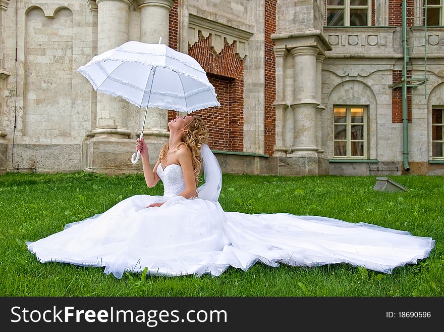 Blonde bride in white dress sitting on the grass at the brick walls of the palace, and looking up at white umbrella - overall plan. Blonde bride in white dress sitting on the grass at the brick walls of the palace, and looking up at white umbrella - overall plan