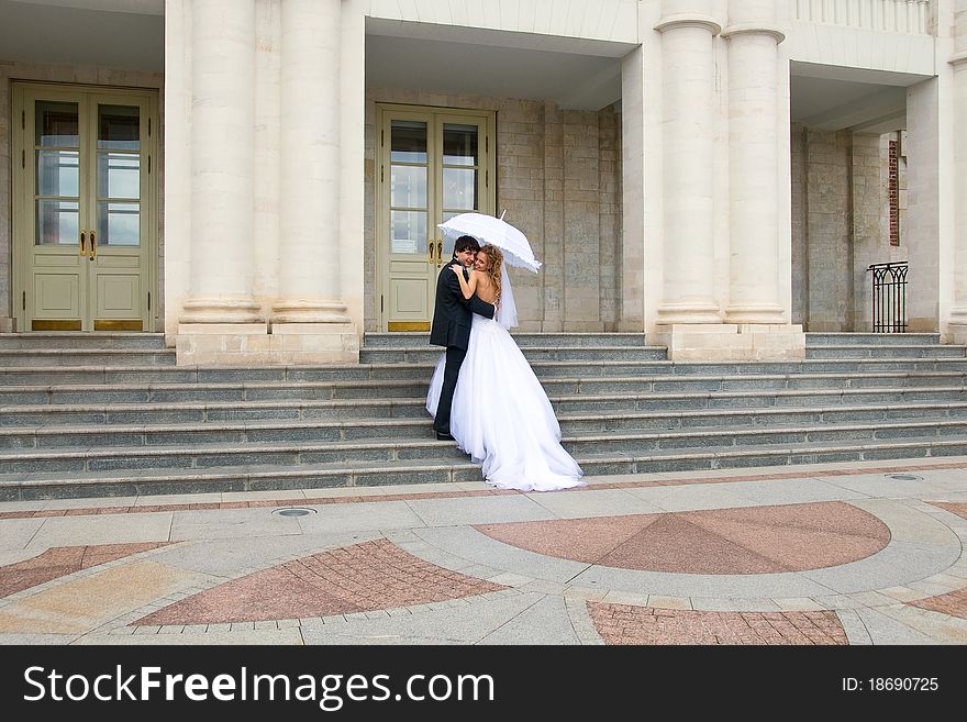 On the steps of the royal palace newlyweds hugging under white umbrella, in the foreground is visible patterned paving. On the steps of the royal palace newlyweds hugging under white umbrella, in the foreground is visible patterned paving