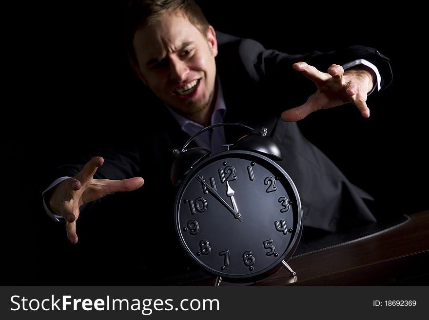 Young desperate businessman in dark suit at office desk jumping at alarm clock showing five minutes to twelve o'clock symbolizing the end is approaching, low-key image isolated on black background. Young desperate businessman in dark suit at office desk jumping at alarm clock showing five minutes to twelve o'clock symbolizing the end is approaching, low-key image isolated on black background.