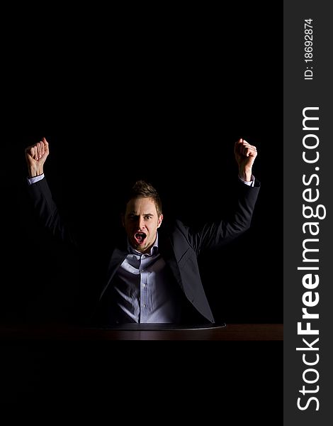 Young modern businessman in dark suit sitting at office desk and raising hands celebrating business success, low-key image isolated on black background. Young modern businessman in dark suit sitting at office desk and raising hands celebrating business success, low-key image isolated on black background.