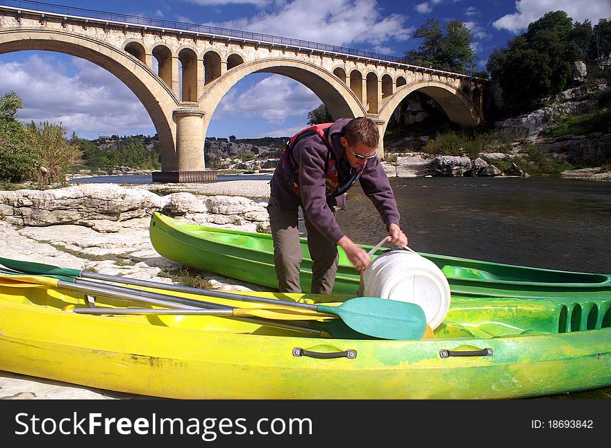 Young man preparing a kayak for a tour on the river Gardon, France, ancient bridge in background. Young man preparing a kayak for a tour on the river Gardon, France, ancient bridge in background