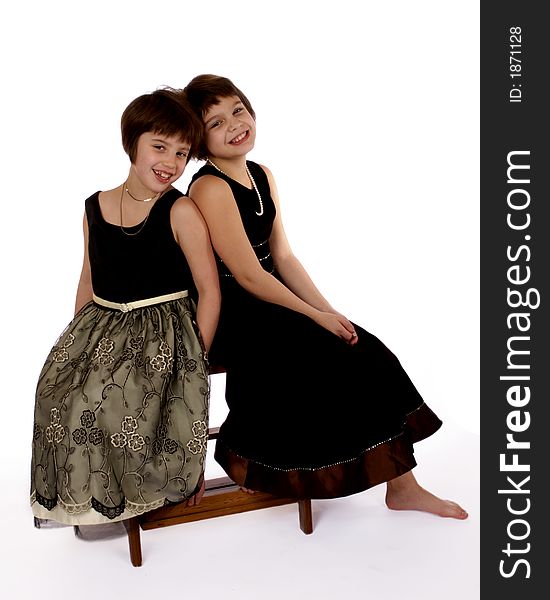 Adorable sisters dressed up for a fancy affair sitting on a stool back to back.   Isolateed on white. Adorable sisters dressed up for a fancy affair sitting on a stool back to back.   Isolateed on white.