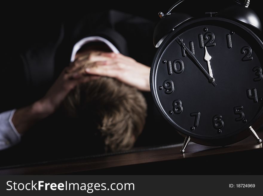 Desperate business person in dark suit sitting at office desk with head down being in despair with close up of alarm clock in foreground showing five minutes to twelve o-c'clock, low-key style isolated on black background. Desperate business person in dark suit sitting at office desk with head down being in despair with close up of alarm clock in foreground showing five minutes to twelve o-c'clock, low-key style isolated on black background.