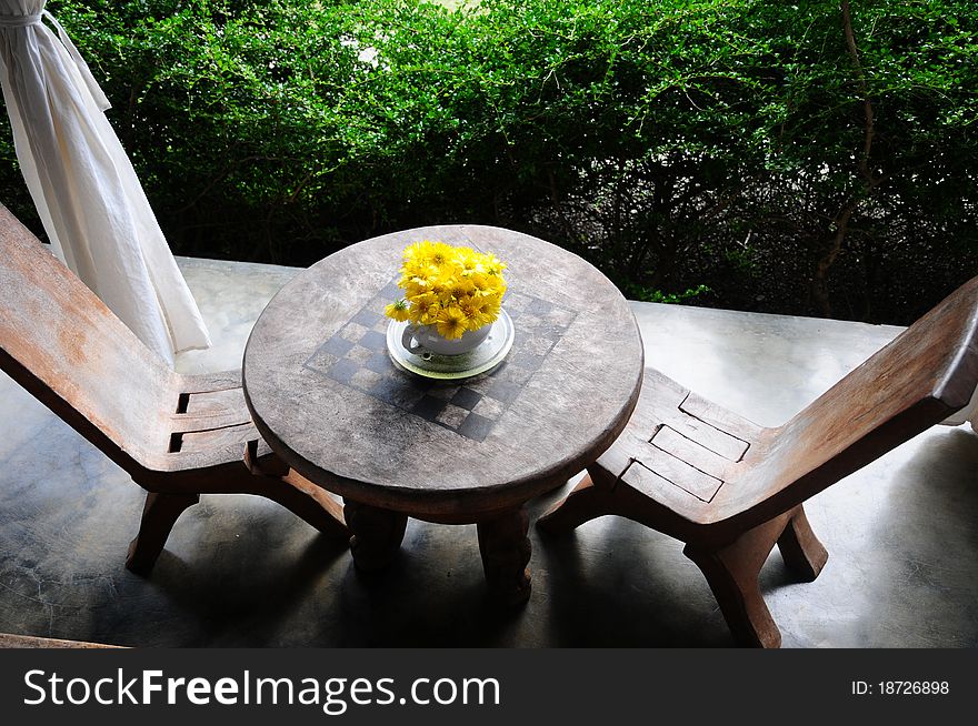 Small sized wooden chairs around a small sized wooden table with chess board imprinted and yellow flowers in a pot. Small sized wooden chairs around a small sized wooden table with chess board imprinted and yellow flowers in a pot.