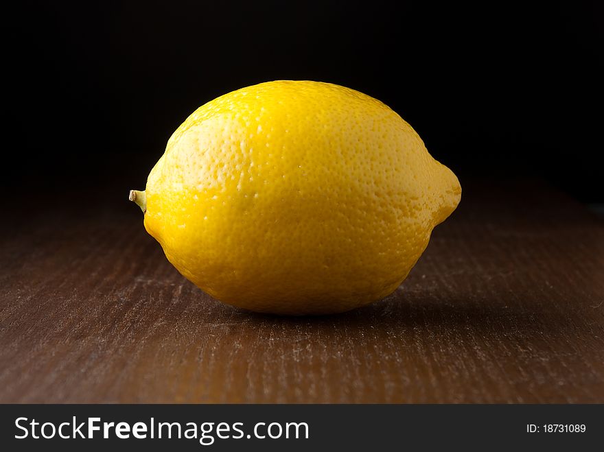 A single fresh yellow lemon on a wood grain table against a black background. A single fresh yellow lemon on a wood grain table against a black background.