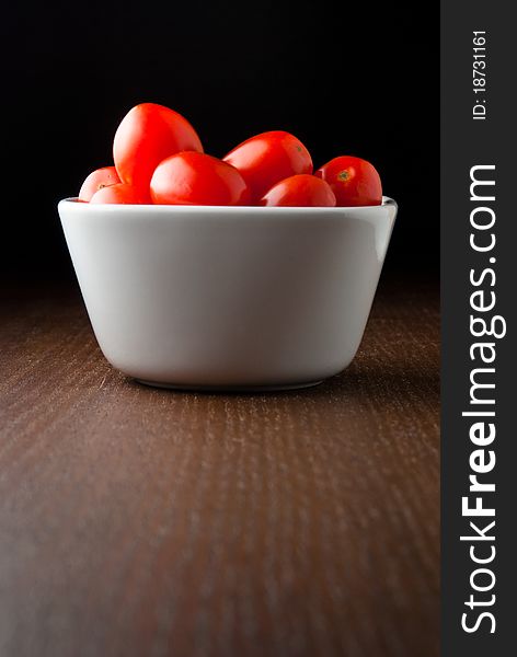 A white bowl of fresh red cherry tomatoes on a wood table against a black background. A white bowl of fresh red cherry tomatoes on a wood table against a black background.