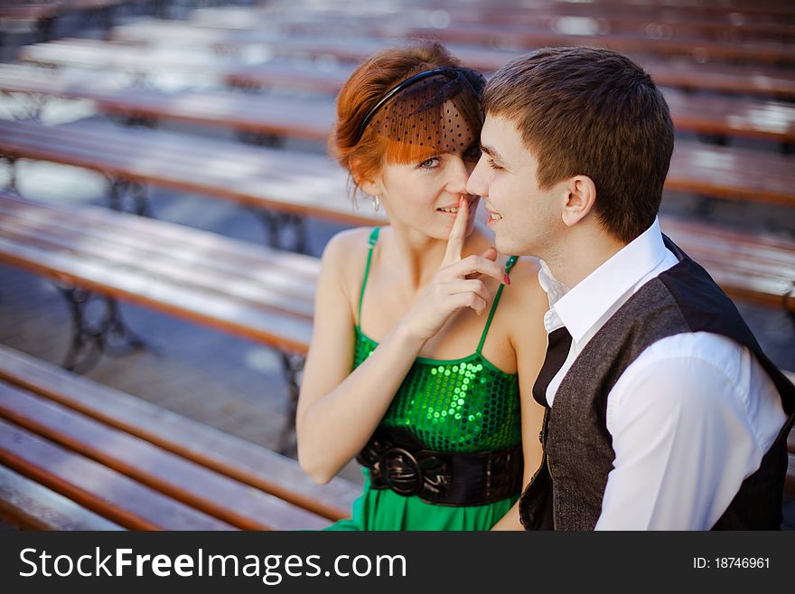 Closeup portraits of young couple sitting together on park bench. Girl making a keep it quiet. Focus on a guy