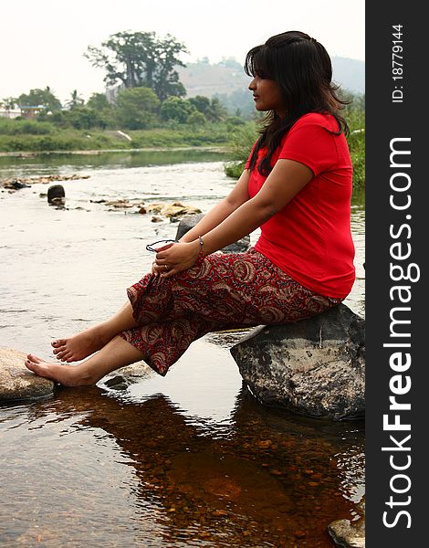 Young indian woman in casual red dress sitting on a rock in water. Young indian woman in casual red dress sitting on a rock in water