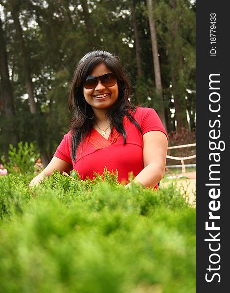 Good looking woman in red dress wearing shades behind waist height bushes. Good looking woman in red dress wearing shades behind waist height bushes.