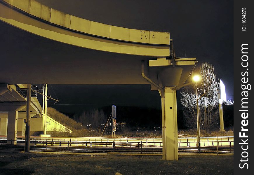 A traffic bridge over tracks at night, with one electric light in front of a tree.