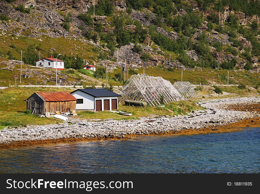 Norwegian shore is photographed in summer. There are some wooden houses and sheds under moss mountain, scaffoldings for dried cod near edge of quite water. Norwegian shore is photographed in summer. There are some wooden houses and sheds under moss mountain, scaffoldings for dried cod near edge of quite water.