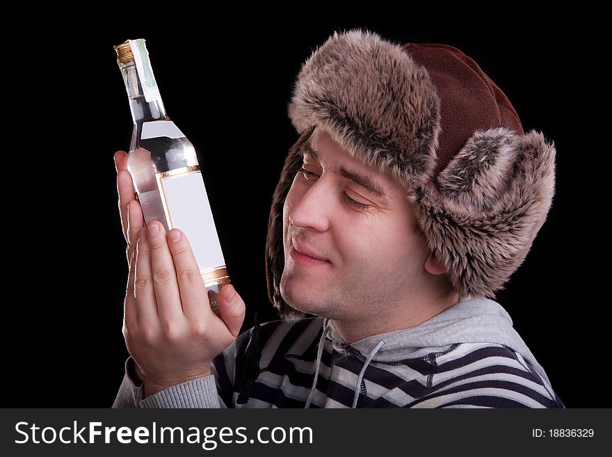 Man in a striped sweater and a fur hat with adoring looks at the bottle of alkohol