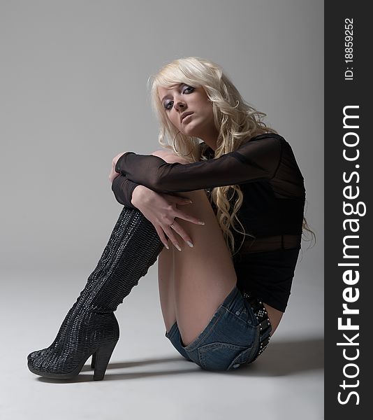 Sexual long-haired blonde girl wearing a jean mini shorts, black shirt and high heels boots posing on the floor in studio on grey background. Sexual long-haired blonde girl wearing a jean mini shorts, black shirt and high heels boots posing on the floor in studio on grey background