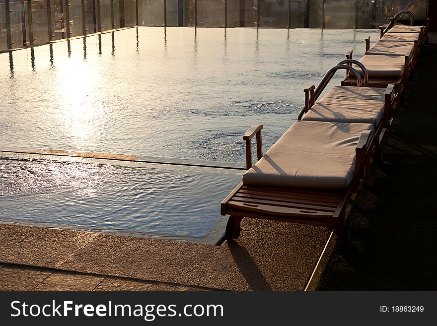 A row of cushioned benches by the side of a hotel pool at sunset. A row of cushioned benches by the side of a hotel pool at sunset.