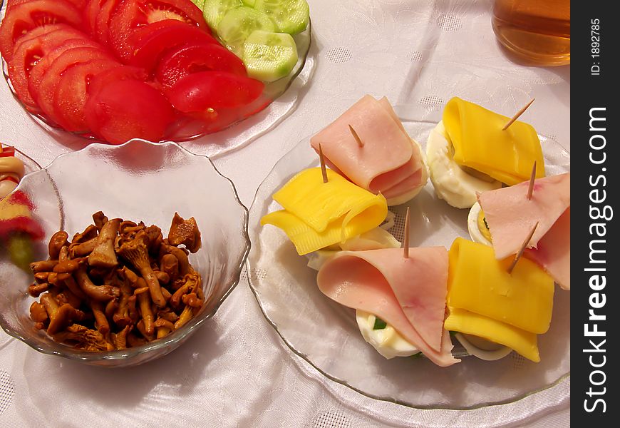 Piece of holiday supper table: tomato, cucumber, pickled cucumbers, cork, red paprika, onion, fungus, egg, cheese, ham, on the plates, all and cup of tea on the tablecloth