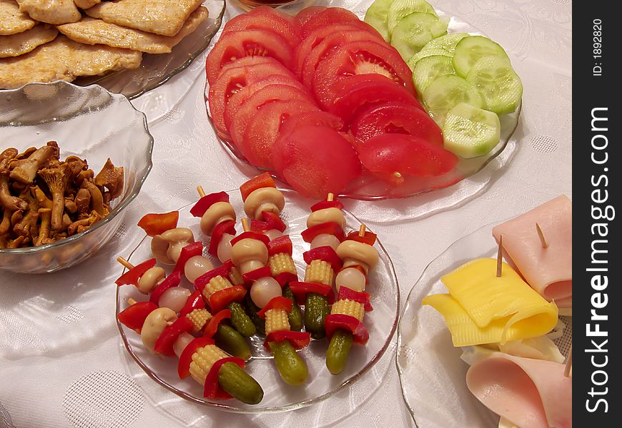 Piece of holiday supper table:
tomato, cucumber, pickled cucumbers, cork, red paprika, onion, fungus, egg, cheese, ham, poultry chop on the plates, 
all on the tablecloth