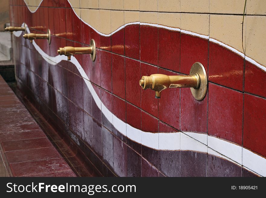 Row of brass water taps over tiled wall; focus on the front tap. Row of brass water taps over tiled wall; focus on the front tap