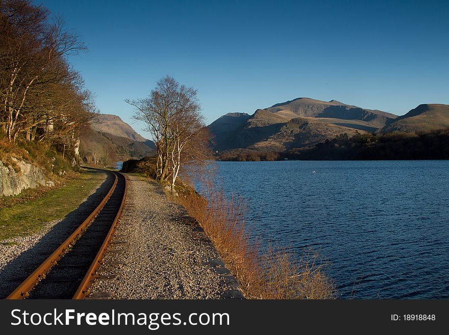A single track rail line next to a lake with trees and mountains in the distance with a blue sky. A single track rail line next to a lake with trees and mountains in the distance with a blue sky.