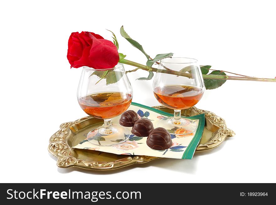 The rose and glasses with cognac and sweets on a tray are isolated on a white background