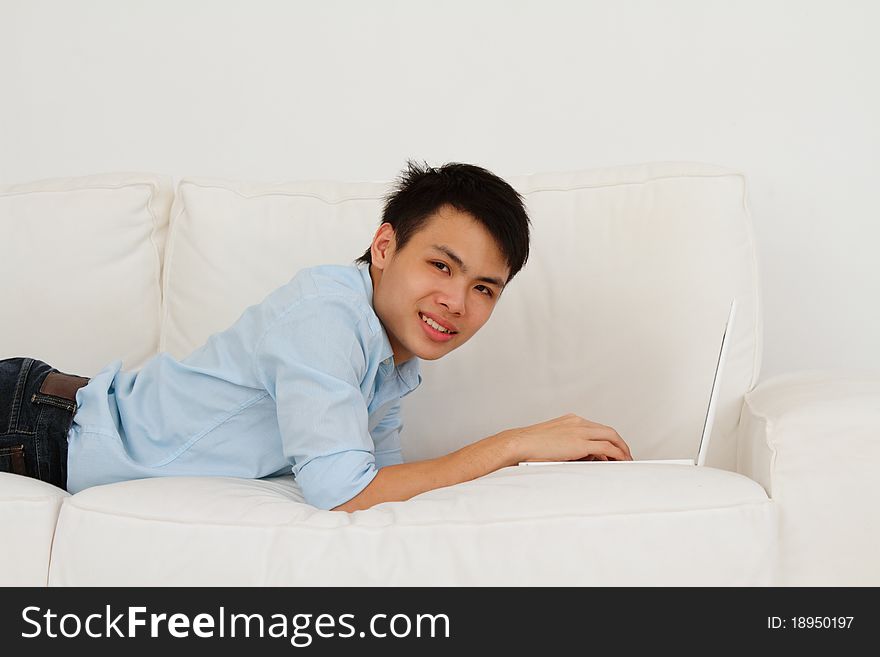 A smiling young man lying prone while using his laptop on a sofa. A smiling young man lying prone while using his laptop on a sofa