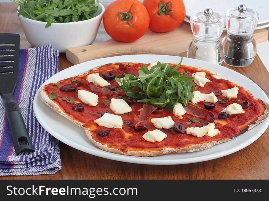 A delicious homemade pizza topped with prosciutto, ricotta cheese, olives and rocket salad. On a wooden kitchen table ready to be served. A delicious homemade pizza topped with prosciutto, ricotta cheese, olives and rocket salad. On a wooden kitchen table ready to be served.