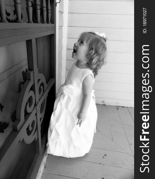 A baby girl is happy while looking at designes on screen door of an old colonial house. She is wearing a white formal dress and white bow. A baby girl is happy while looking at designes on screen door of an old colonial house. She is wearing a white formal dress and white bow.