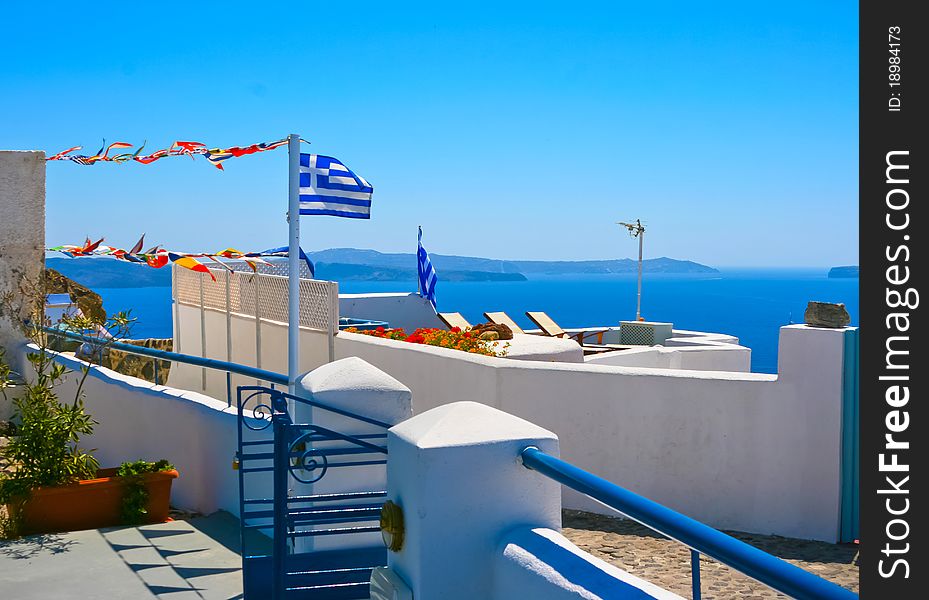 Beautiful white terrace at resort on island of Santorini in Oia, Greece. Traditional greek architecture and national flag of Greece. Aegean sea in a background. Beautiful white terrace at resort on island of Santorini in Oia, Greece. Traditional greek architecture and national flag of Greece. Aegean sea in a background.