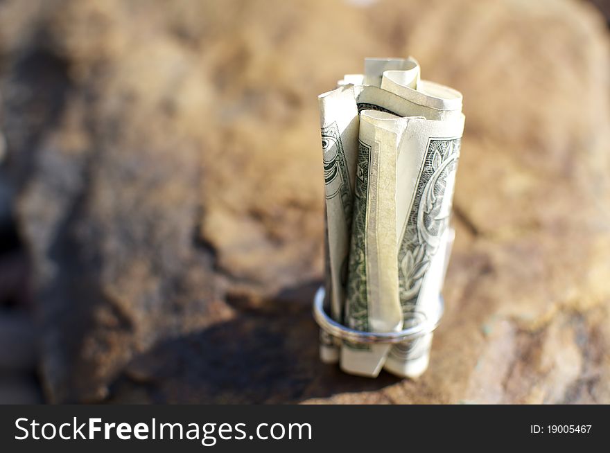 A US dollar is rolled up and sits on a rock outside. A US dollar is rolled up and sits on a rock outside.