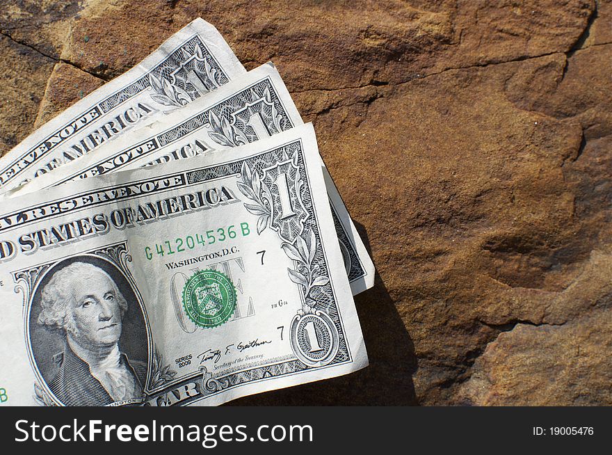 Three one dollar bills sit on a rock waiting for someone to find them.