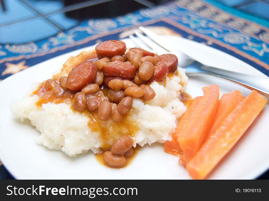 Franks and beans over mashed potatoes, with cooked carrots in a white plate on a place setting, with fork.