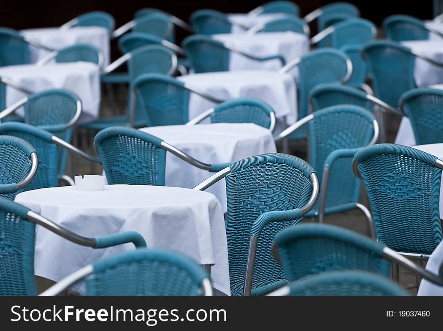 Many empty blue chairs with white rounded tables outside in front of restaurant. Many empty blue chairs with white rounded tables outside in front of restaurant.