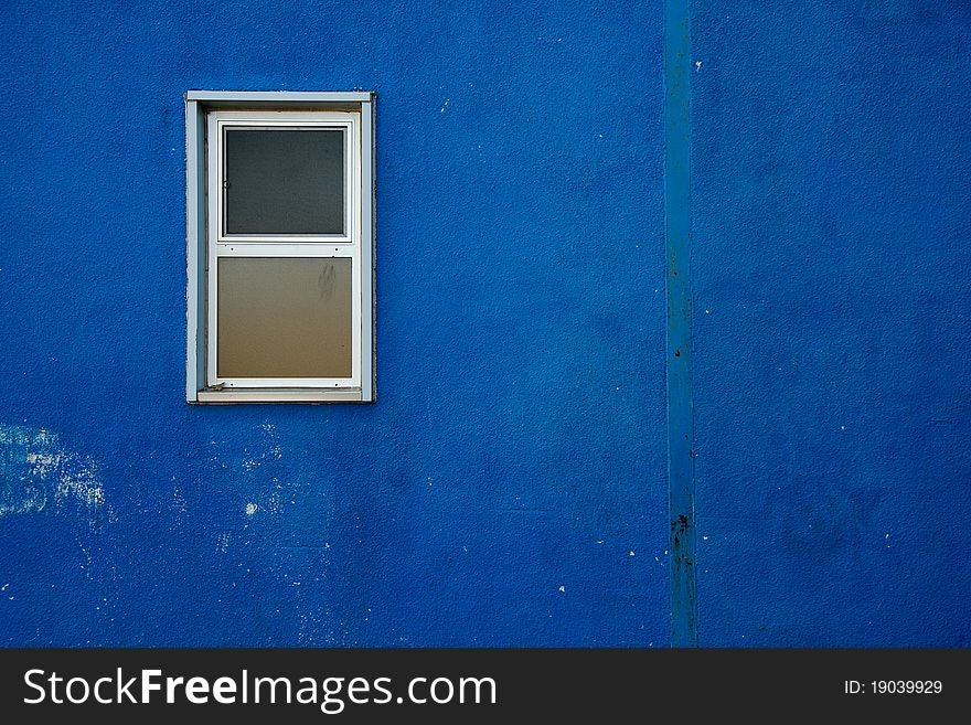 A small window on a blue, rough and worn exterior wall. A small window on a blue, rough and worn exterior wall.