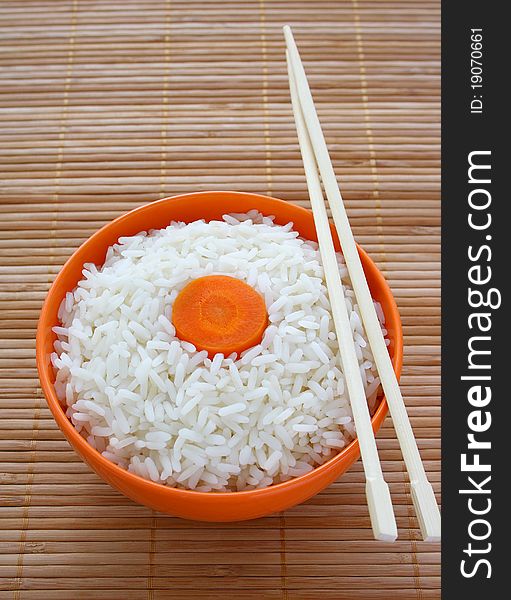 Orange bowl of cooked rice decorated with carrots and sticks on bamboo napkin. Orange bowl of cooked rice decorated with carrots and sticks on bamboo napkin.