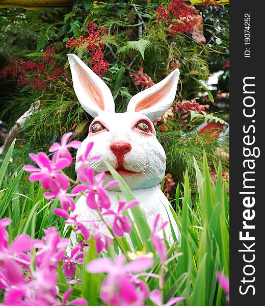 A statue of a white, pink-nosed rabbit sitting in a garden surrounded by flowers. A statue of a white, pink-nosed rabbit sitting in a garden surrounded by flowers