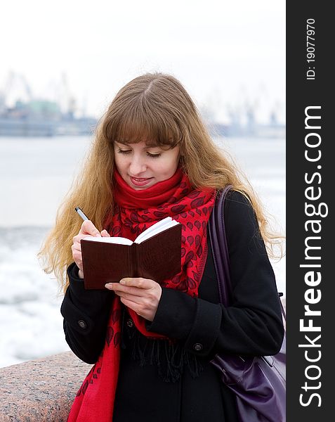 Beautiful young girl in the red scarf and black coat writing in notepad. Beautiful young girl in the red scarf and black coat writing in notepad