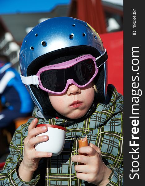 Little girl in ski helmet and jacket eats sausage and drink tea. Little girl in ski helmet and jacket eats sausage and drink tea