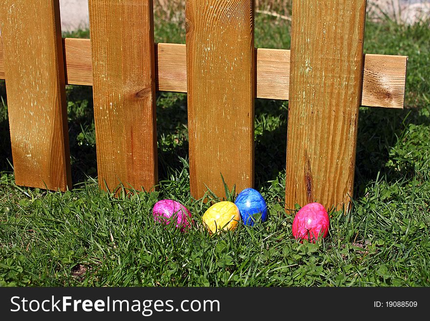 Some coloured easter eggs lying in the grass in front of a wooden fence