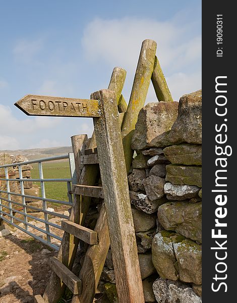 Wooden footpath sign and stile across stone wall in the sunshine beside metal gate