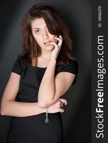 Portrait of beautiful and young woman in black dress with dark hair. Looking into the camera, hand at her face, dark background. Portrait of beautiful and young woman in black dress with dark hair. Looking into the camera, hand at her face, dark background.