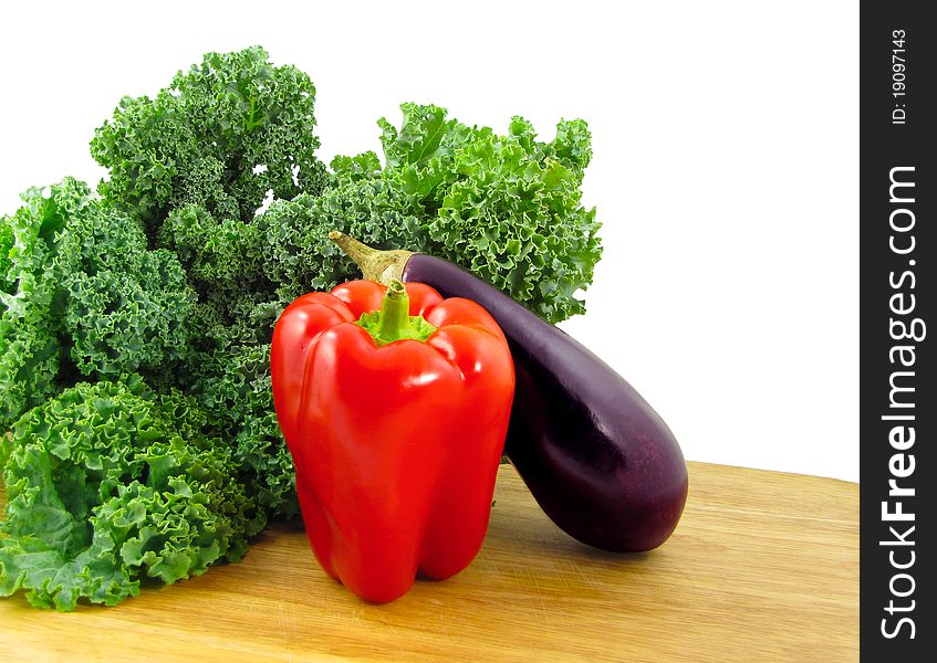 A red bell pepper, a purple eggplant, and a bunch of green kale on a wooden cutting board isolated on white. A red bell pepper, a purple eggplant, and a bunch of green kale on a wooden cutting board isolated on white.