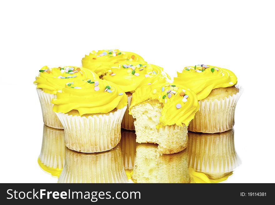 Several white cup cakes with yellow frosting and sprinkles with one of them having a bite taken out of it. Several white cup cakes with yellow frosting and sprinkles with one of them having a bite taken out of it
