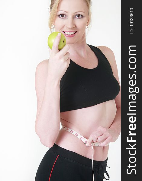 Attractive blond caucasian woman smiling wearing fitness attire holding an apple with measuring tape around thin waist. Attractive blond caucasian woman smiling wearing fitness attire holding an apple with measuring tape around thin waist