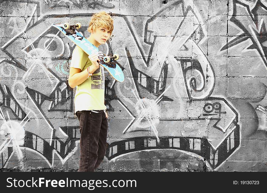 A boy with a skateboard in an urban setting with black and white graffiti in the background. A boy with a skateboard in an urban setting with black and white graffiti in the background