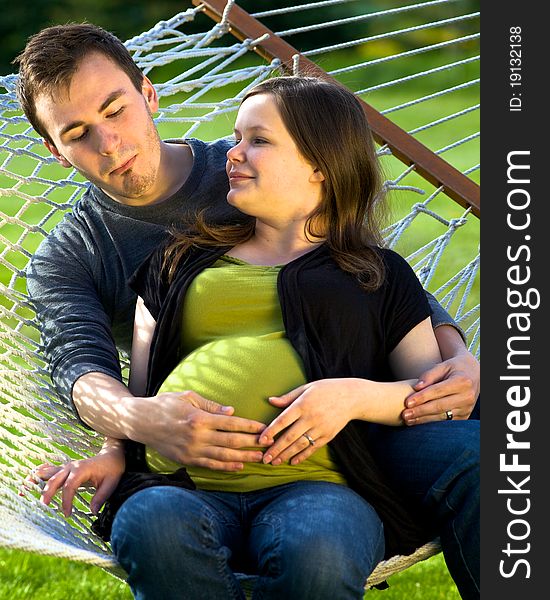 Young man sits on a hammock admiring his wifeâ€™s pregnant belly by making a heart with his hands. Young man sits on a hammock admiring his wifeâ€™s pregnant belly by making a heart with his hands.