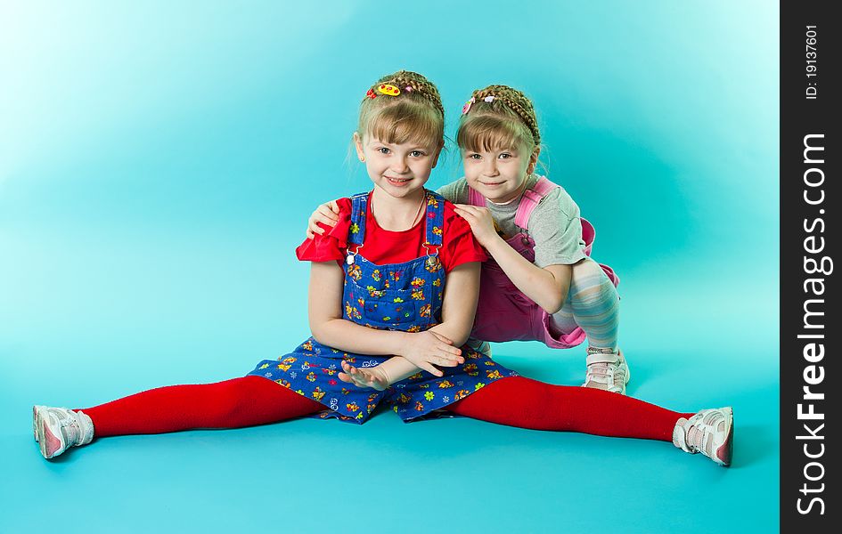 A girl trying to do the splits exercise, her twin sister sitting besides her over blue background. A girl trying to do the splits exercise, her twin sister sitting besides her over blue background